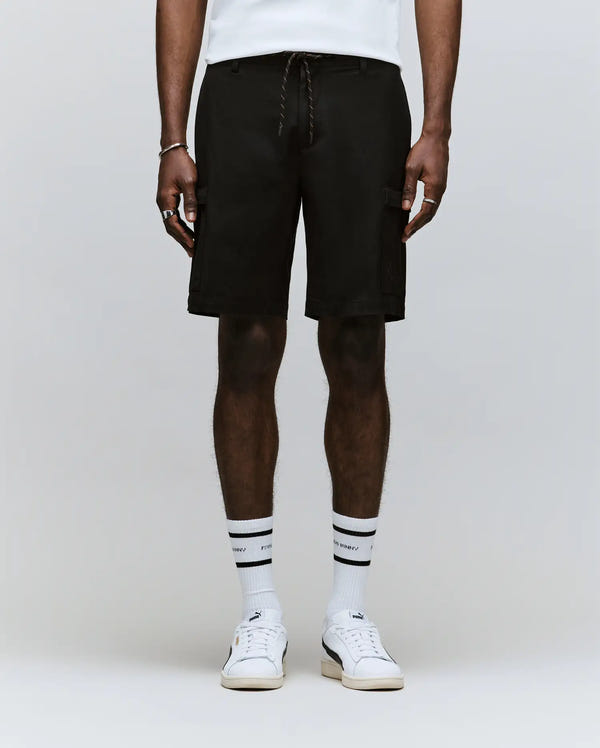 A person wears Psycho Bunny MENS ARLO CARGO SHORT - MB0700007, black with zippered cargo pockets, plus white athletic socks with black stripes and white sneakers. Only the body from waist to feet is visible against a plain light background.