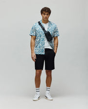Relaxed fit short-sleeve shirt in white with an all-over teal pixelated Psycho Bunny skull logo pattern, layered over a white crew neck tee, paired with classic black Mens Shiro Twill Short.