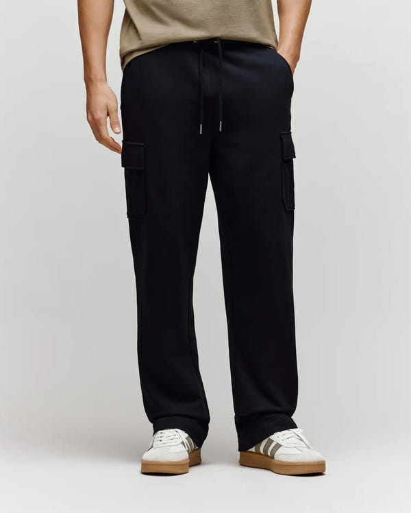 A person wearing Psycho Bunny MENS NASH CARGO SWEATPANT - MB0800010, white sneakers with brown soles, and a beige shirt stands against a plain light background.