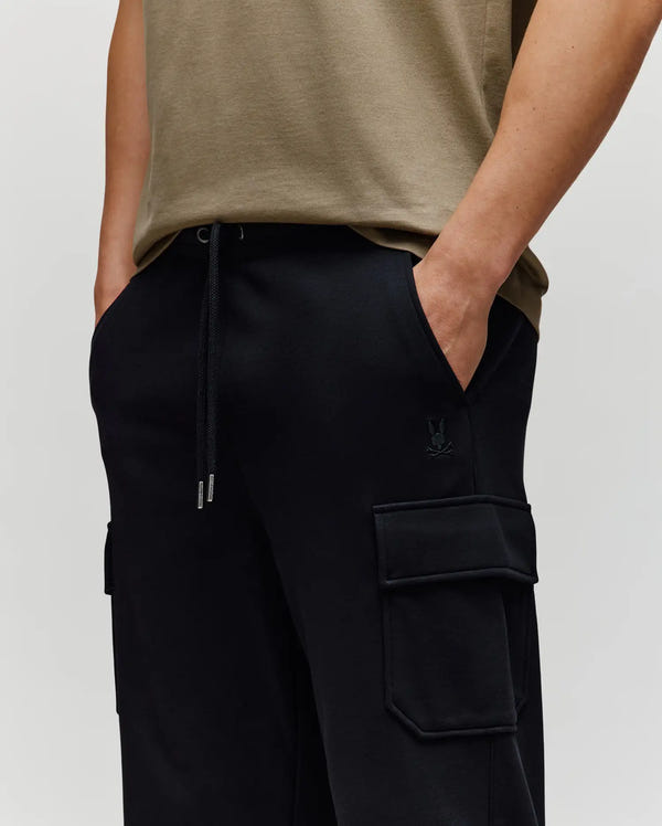A person stands hands in pockets against a plain background, showcasing effortless style and comfort in Psycho Bunny’s MENS NASH CARGO SWEATPANT - MB0800010, featuring black cargo sweatpants with side cargo pockets.