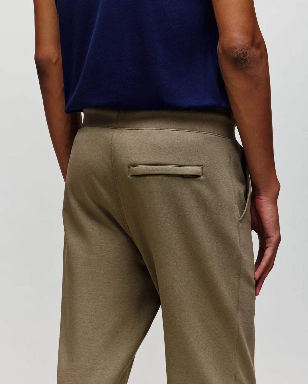A person is seen from the back wearing Psycho Bunny MENS WADE SWEATPANT - MB0800014 in olive green with a classic athletic fit and single back welt pocket, paired with a navy blue shirt. One hand rests at their side.