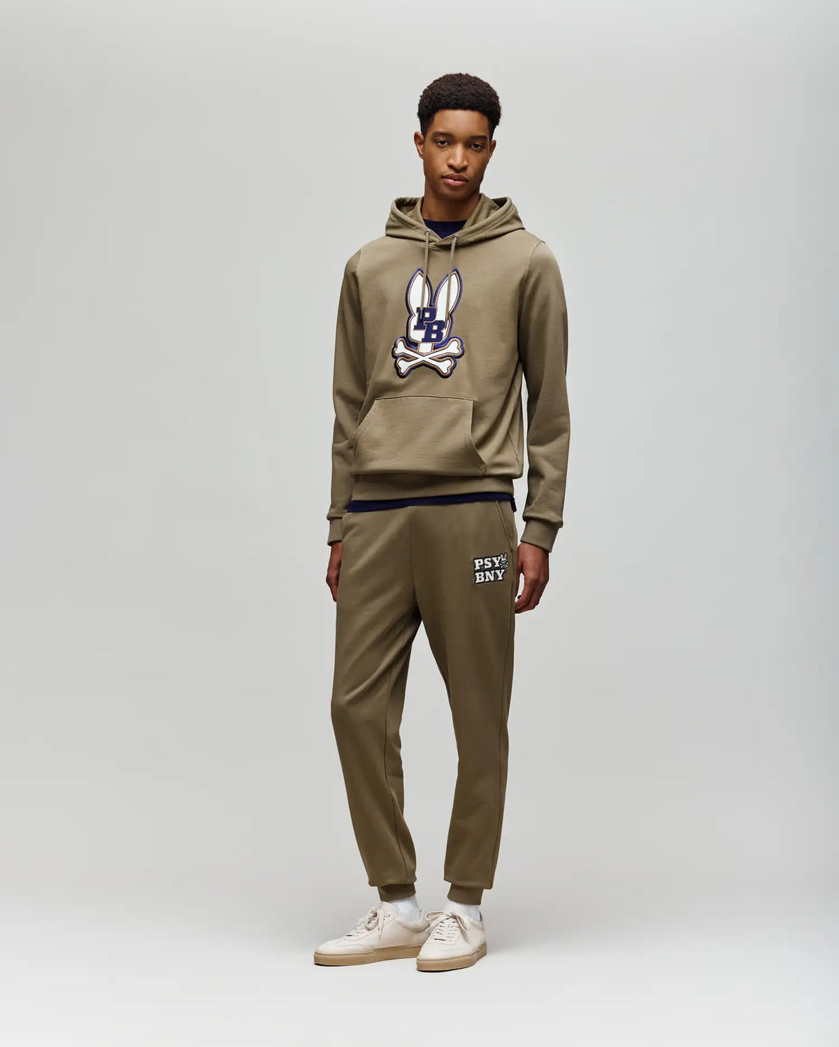 A young man stands against a light gray background, wearing Psycho Bunny's olive green hoodie and matching MENS WADE SWEATPANT - MB0800014, both crafted from a premium cotton blend, paired with white sneakers for a classic athletic style.