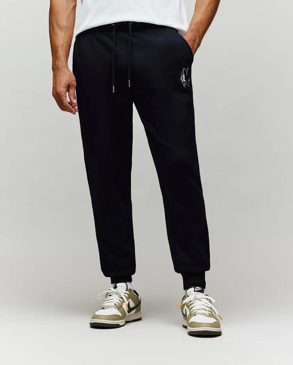 A person stands against a plain light background wearing Psycho Bunny MENS AUGUSTE JOGGER - MB0800015 in black with a small chenille patch on the left thigh, paired with a white T-shirt and beige and white sneakers. Made from 100% cotton for all-day comfort.