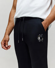 A person wears Psycho Bunny MENS AUGUSTE JOGGER - MB0800015 in black, featuring a small embroidered cartoon bunny and crossbones chenille patch on the pocket, paired with a white T-shirt. Only the torso and upper legs are visible.