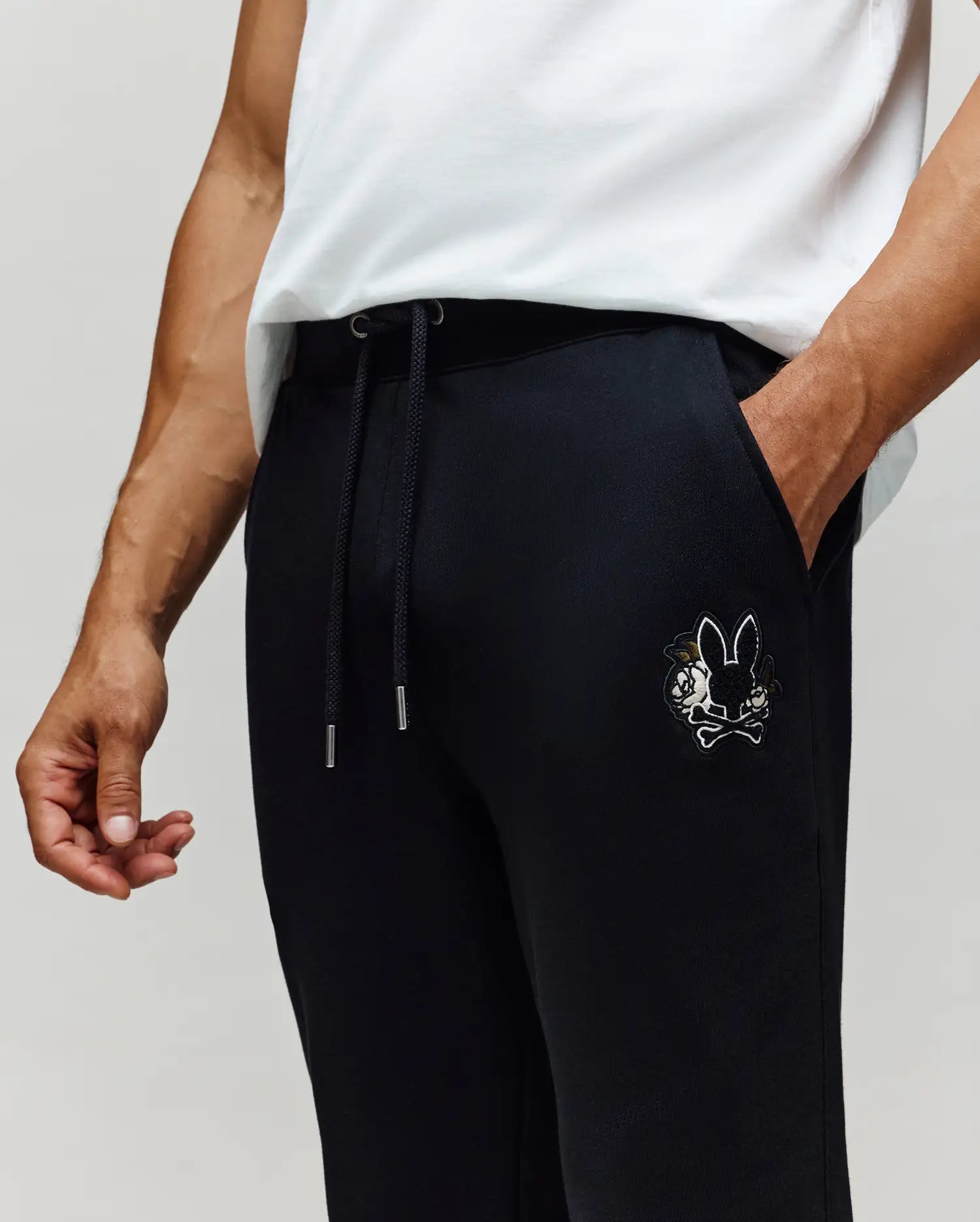 A person wears Psycho Bunny MENS AUGUSTE JOGGER - MB0800015 in black, featuring a small embroidered cartoon bunny and crossbones chenille patch on the pocket, paired with a white T-shirt. Only the torso and upper legs are visible.