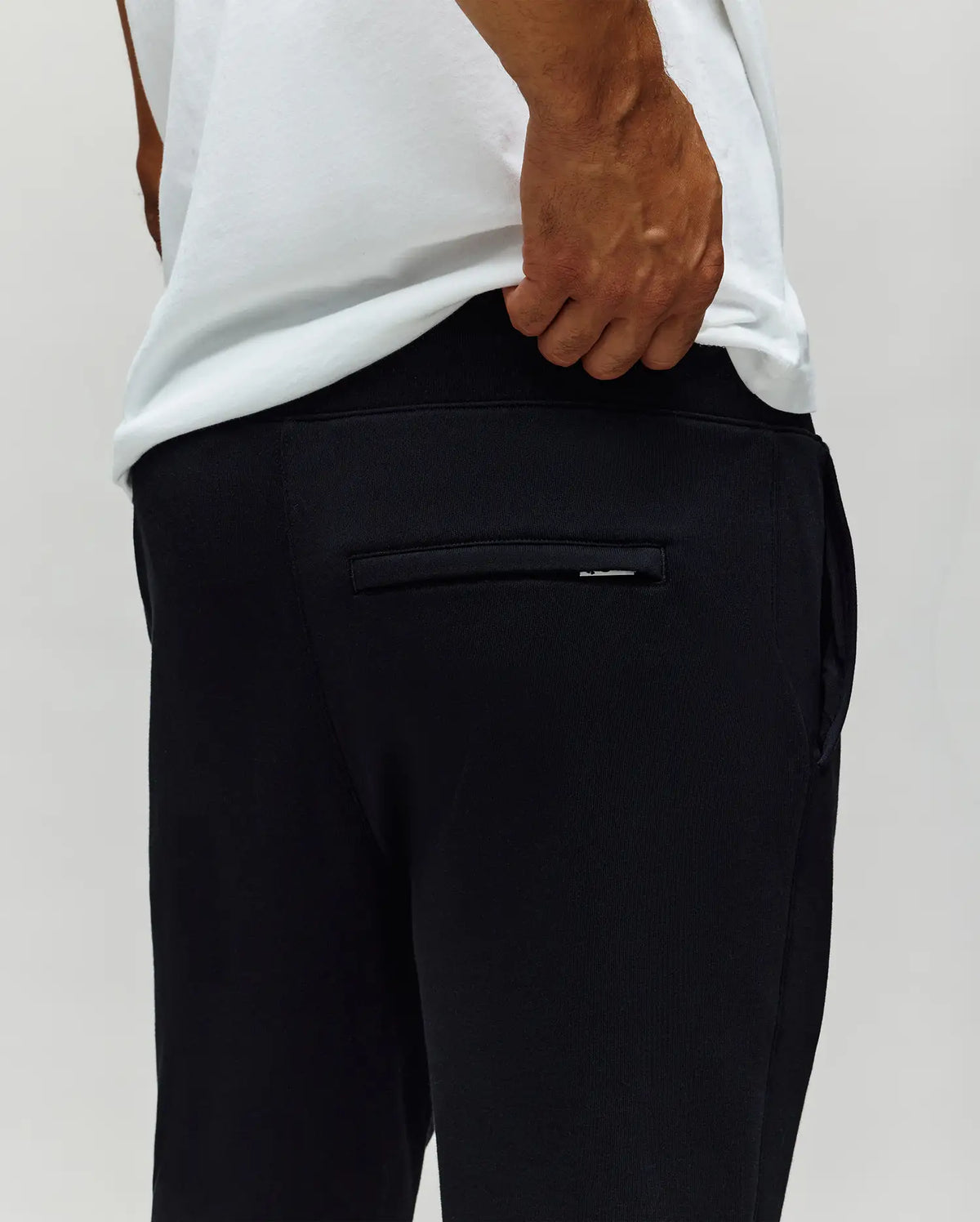 A person wearing a white t-shirt and Psycho Bunny MENS AUGUSTE JOGGER - MB0800015 in black 100% cotton stands with a hand near the waistband, showing off the back pocket detail against a plain light background.