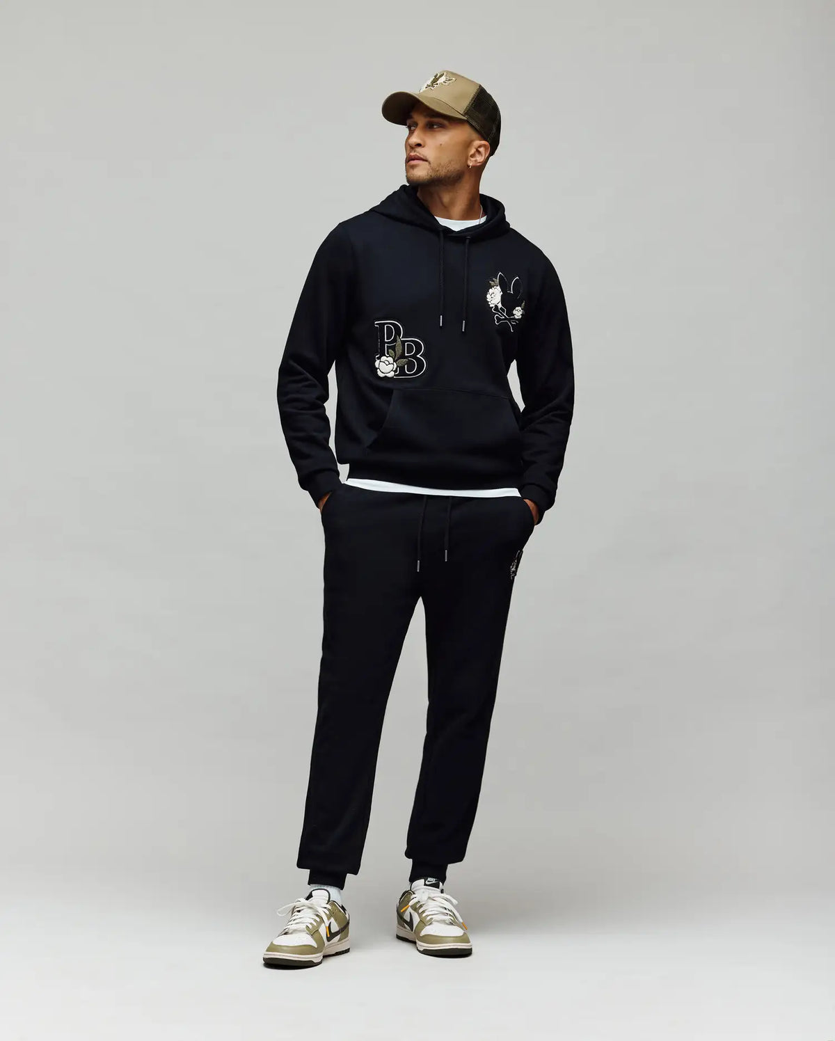 A man in Psycho Bunny MENS AUGUSTE JOGGER - MB0800015 with chenille patches, a black hoodie with white graphics, a tan cap, and white sneakers with gold accents stands against a gray background looking to the side.