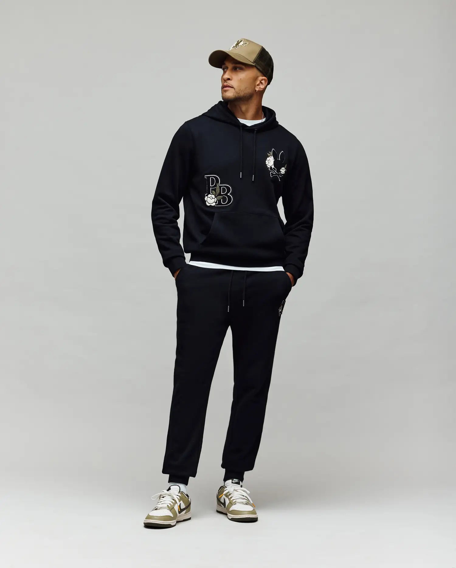 A man in Psycho Bunny MENS AUGUSTE JOGGER - MB0800015 with chenille patches, a black hoodie with white graphics, a tan cap, and white sneakers with gold accents stands against a gray background looking to the side.