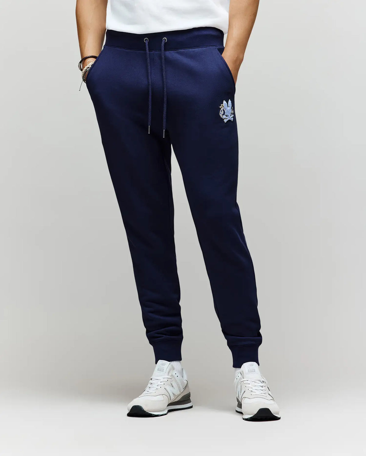A person wears Psycho Bunny MENS AUGUSTE JOGGER - MB0800015 in navy blue, crafted from 100% cotton with a small embroidered crest on the left thigh, paired with white sneakers and a white shirt against a plain, light background.