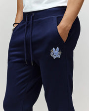 Wearing Psycho Bunny MENS AUGUSTE JOGGER - MB0800015 in navy blue, featuring a blue chenille patch on the left thigh. Made from 100% cotton for comfort and style, paired with hands in pockets and a white shirt partly visible.