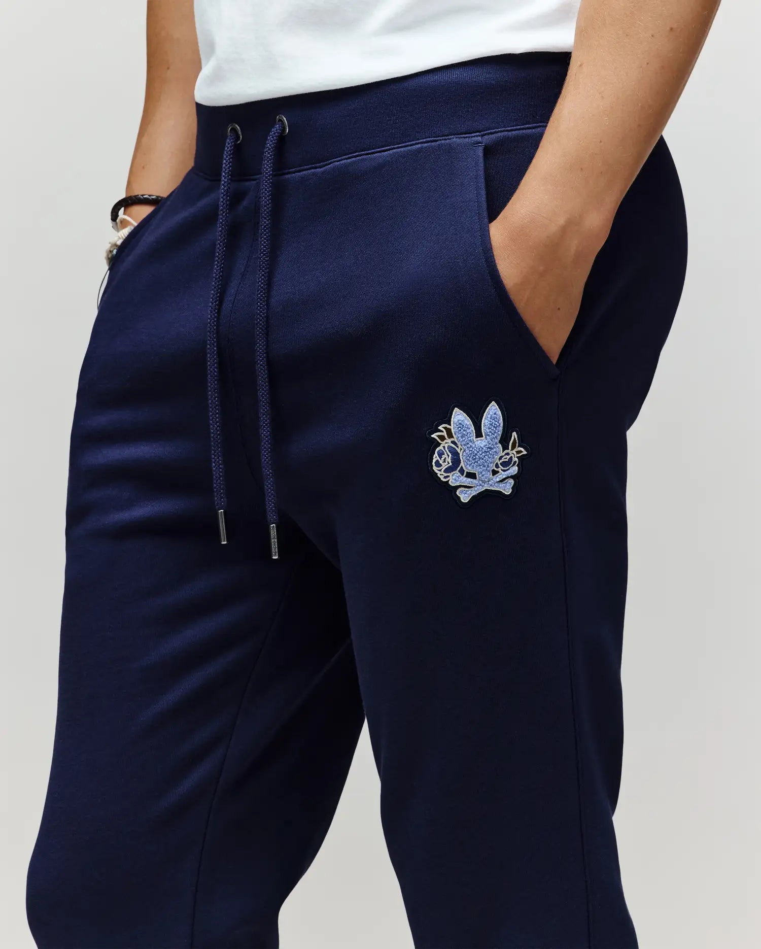 Wearing Psycho Bunny MENS AUGUSTE JOGGER - MB0800015 in navy blue, featuring a blue chenille patch on the left thigh. Made from 100% cotton for comfort and style, paired with hands in pockets and a white shirt partly visible.