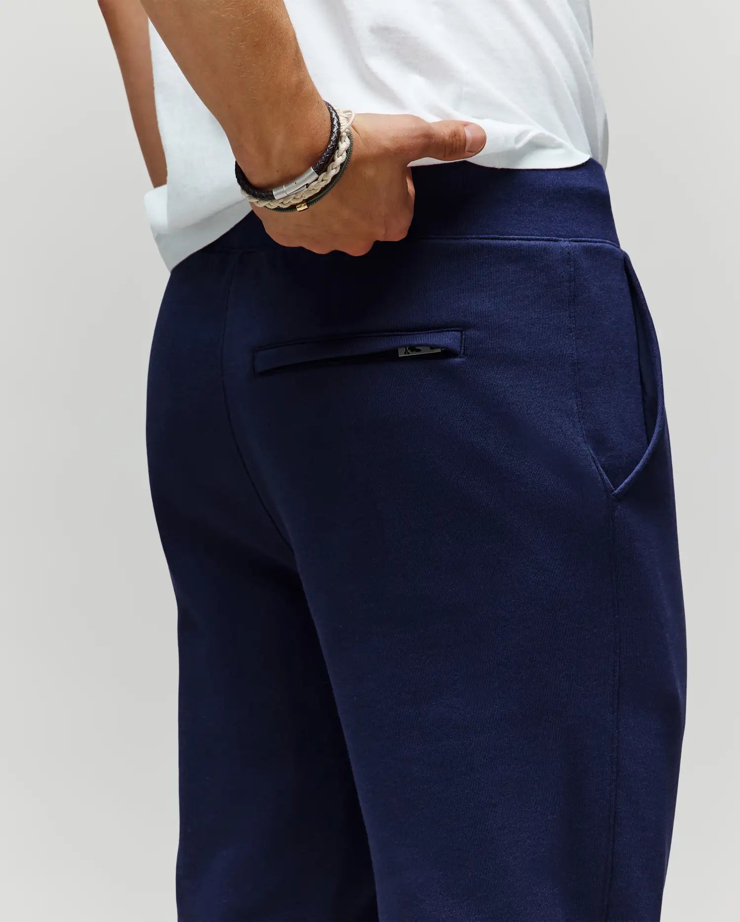 A person wears Psycho Bunny MENS AUGUSTE JOGGER - MB0800015 in navy blue (100% cotton) with a white shirt, their right hand in the zippered back pocket, showing the zipper detail and bracelets on their wrist.