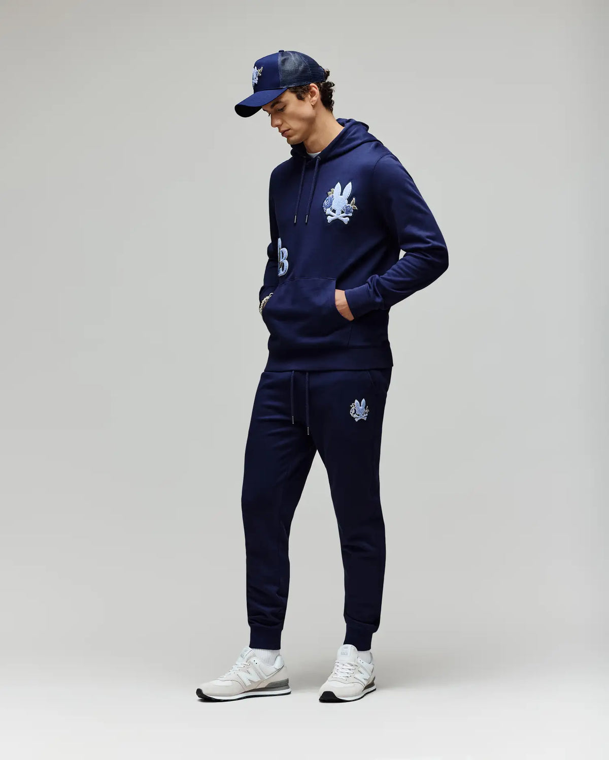 A man in Psycho Bunny MENS AUGUSTE JOGGER - MB0800015, a navy hoodie with cartoon graphics and chenille patches, a matching cap, and white sneakers stands with hands in pockets against a light background.
