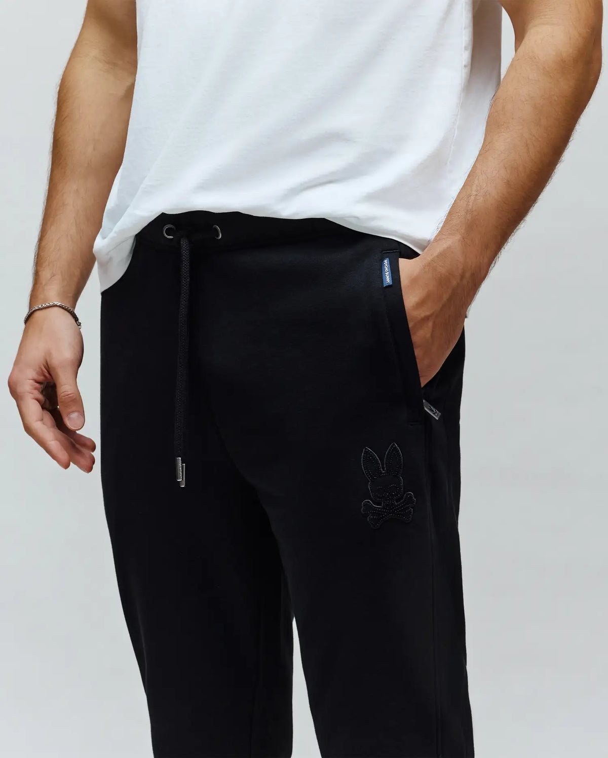 Mens Barry Joggers, black relaxed fit cotton blend joggers with drawstring waist, metal tips, side pockets, large tonal embroidered Psycho Bunny skull logo, and small woven brand tag.