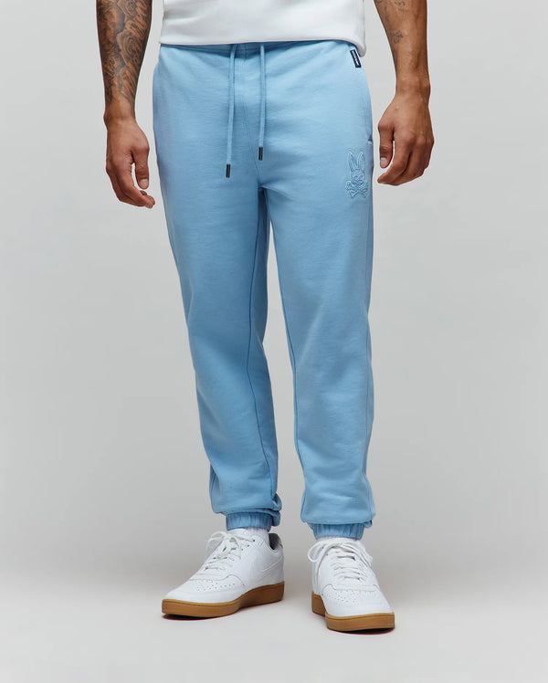 Mens Barry Joggers in light blue, featuring a relaxed fit, soft jersey knit, drawstring waist with black tips, cuffed ankles, and an embroidered Psycho Bunny skull logo on the left thigh.