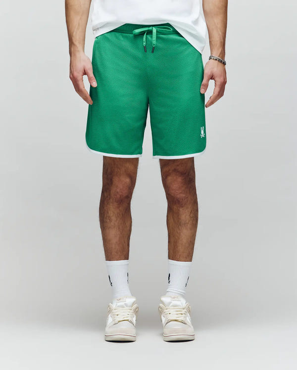 A person wearing Psycho Bunny MENS NICO MESH SHORT - MB0900004 in green with white trim, a white T-shirt, white crew socks, and white sneakers stands against a light gray background.