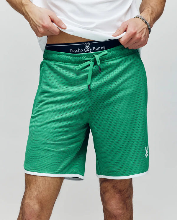 A person wears Psycho Bunny MENS NICO MESH SHORT - MB0900004 in green with white trim and logo, styled with a white t-shirt. The breathable mesh shorts have a drawstring waist, and the person is slightly lifting their shirt.