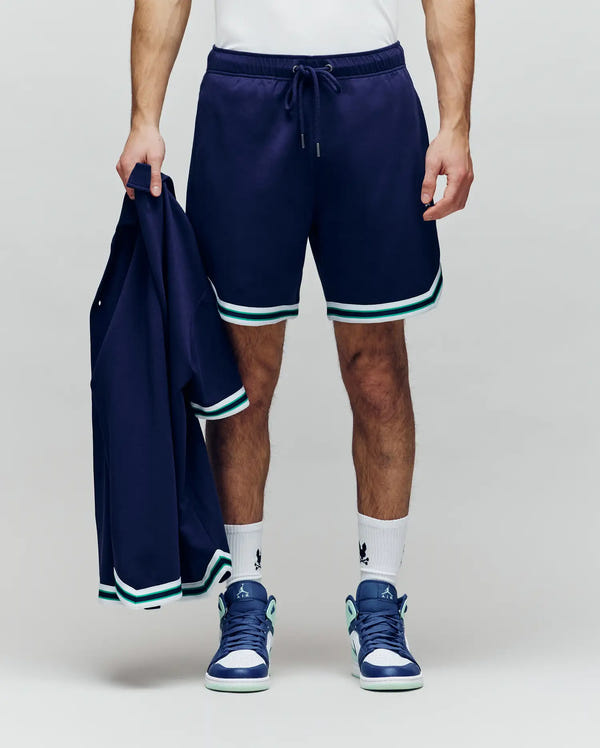 A person wearing Psycho Bunny MENS BASIL TAPED SHORT - MB0900005 with an elastic waistband, white socks, and blue sneakers holds a matching navy jacket against a plain light background; their upper body is partly out of frame.