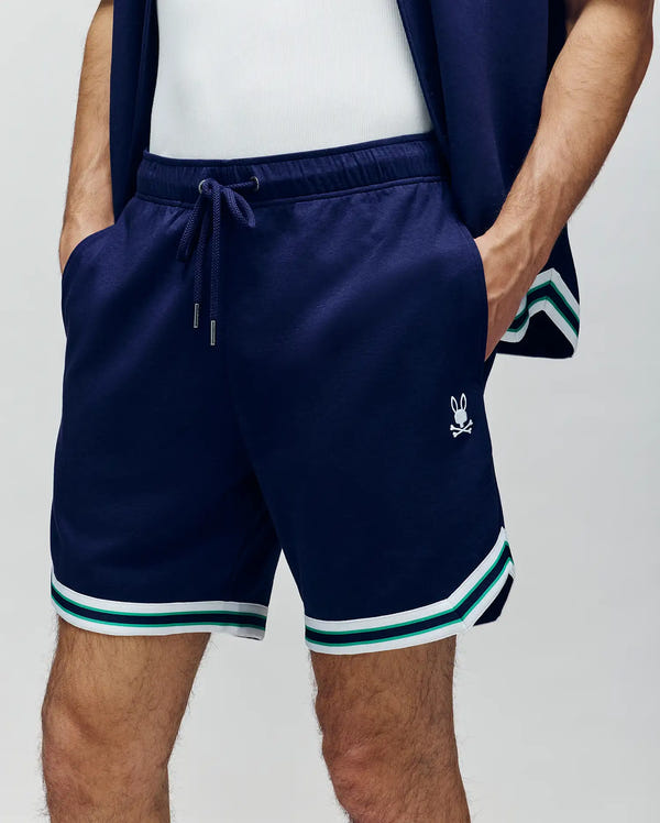 A person wearing Psycho Bunny MENS BASIL TAPED SHORT - MB0900005 in navy blue, featuring an elastic waistband, white and green striped trim, a small bunny logo on the left leg, and hands in pockets.