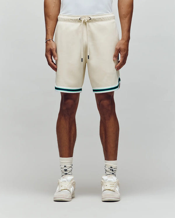 A person wearing Psycho Bunny’s MENS BASIL TAPED SHORT - MB0900005 in cream with teal and black trim, paired with white socks, white sneakers, and a white shirt stands against a plain light gray background.