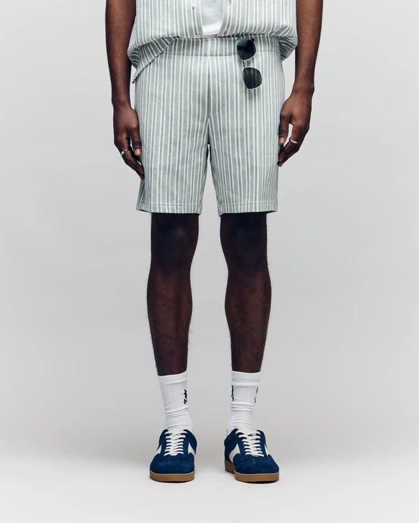 A person wears Psycho Bunny MENS ROBERT KNIT STRIPE SHORT - MB0900007 in light gray and white, paired with white socks, blue sneakers, and sunglasses hanging from the shorts pocket, standing against a plain background.