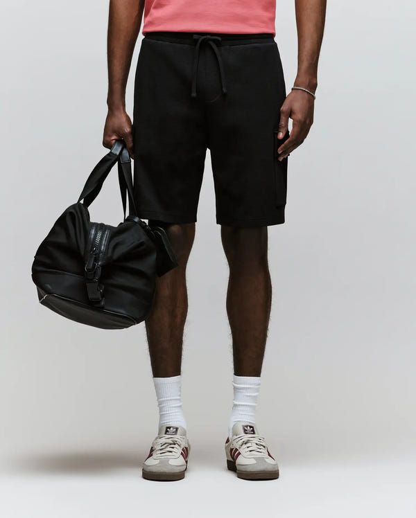 A person stands against a plain background wearing Psycho Bunny’s MENS RON WAFFLE CARGO SHORT - MB0900012 in black, a pink shirt, white socks, and white sneakers, holding a black duffel bag in their left hand. Their face is not visible.