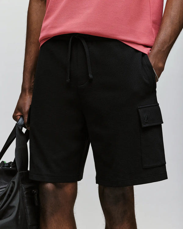 A person wears Psycho Bunny’s MENS RON WAFFLE CARGO SHORT - MB0900012, featuring a drawstring waist, large pocket, and embroidered logos. Paired with a pink shirt and holding a black bag; only the lower torso and upper legs are shown.
