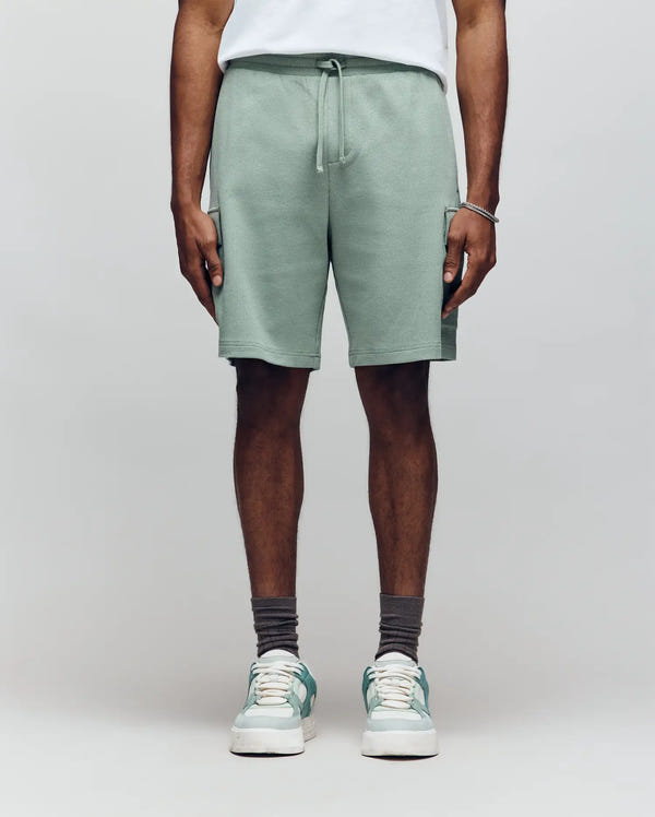 A person wears Psycho Bunny MENS RON WAFFLE CARGO SHORT - MB0900012 in sage green, paired with a white tee, gray socks, and white sneakers with green accents. Photo shows only the body from shoulders down against a plain light background.