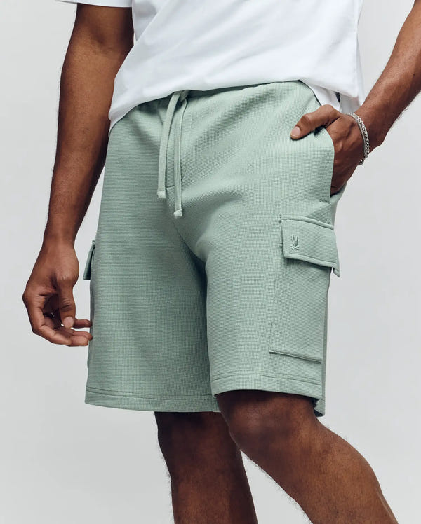 Wearing Psycho Bunny's MENS RON WAFFLE CARGO SHORT - MB0900012 in light green with flap pockets and a drawstring waist, paired with a white t-shirt accented by subtle embroidered logos and accessorized with a bracelet.