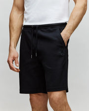 A person in a white t-shirt and Psycho Bunny MENS KAI SWEATSHORT - MB0900015, black with twill texture and an embroidered Bunny logo, stands with one hand in their pocket against a plain light background.