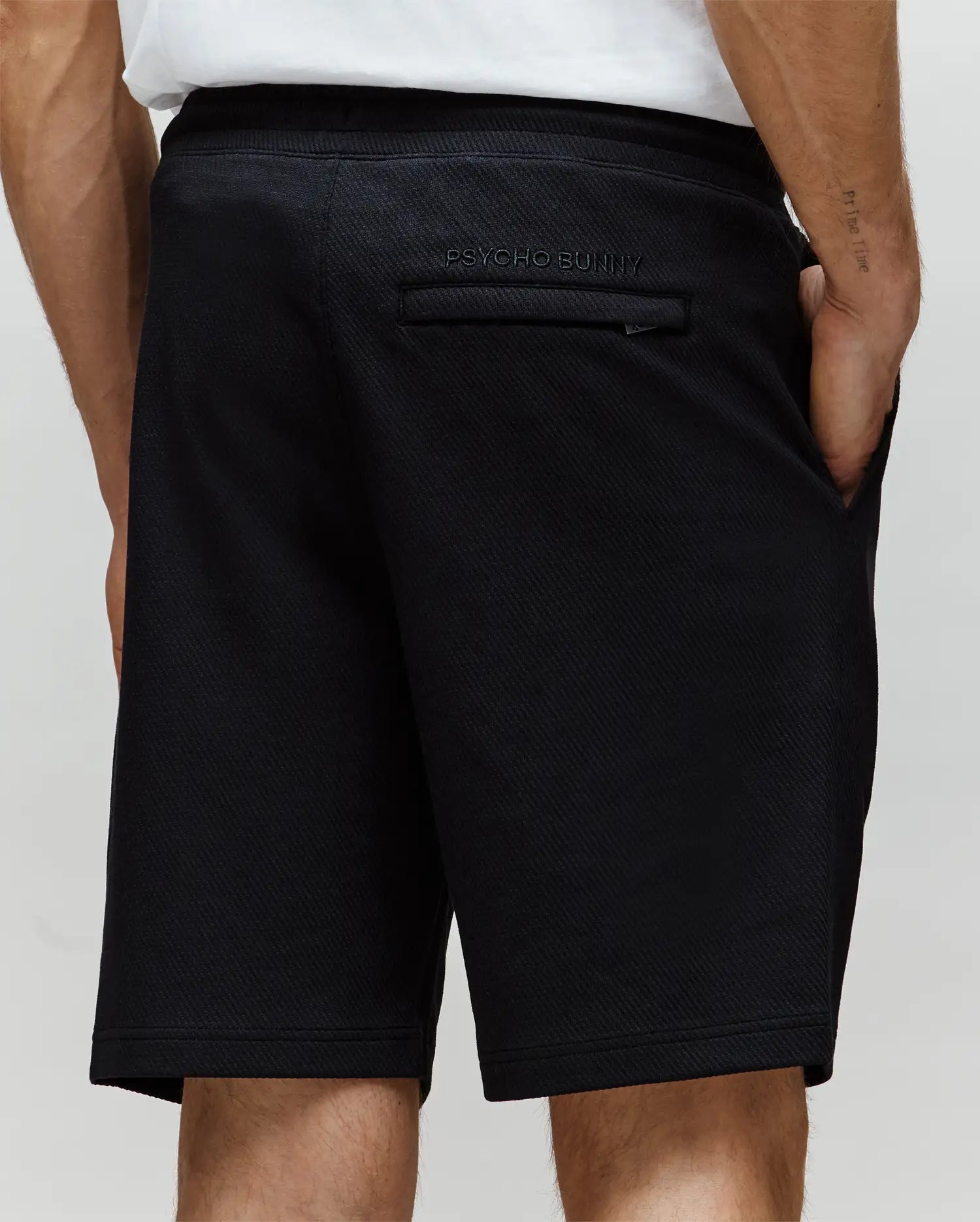 A person wears black MENS KAI SWEATSHORT - MB0900015 by Psycho Bunny, featuring twill texture, one hand in the pocket, and a back pocket with "Psycho Bunny" text above an embroidered Bunny logo, paired with a crisp white shirt.