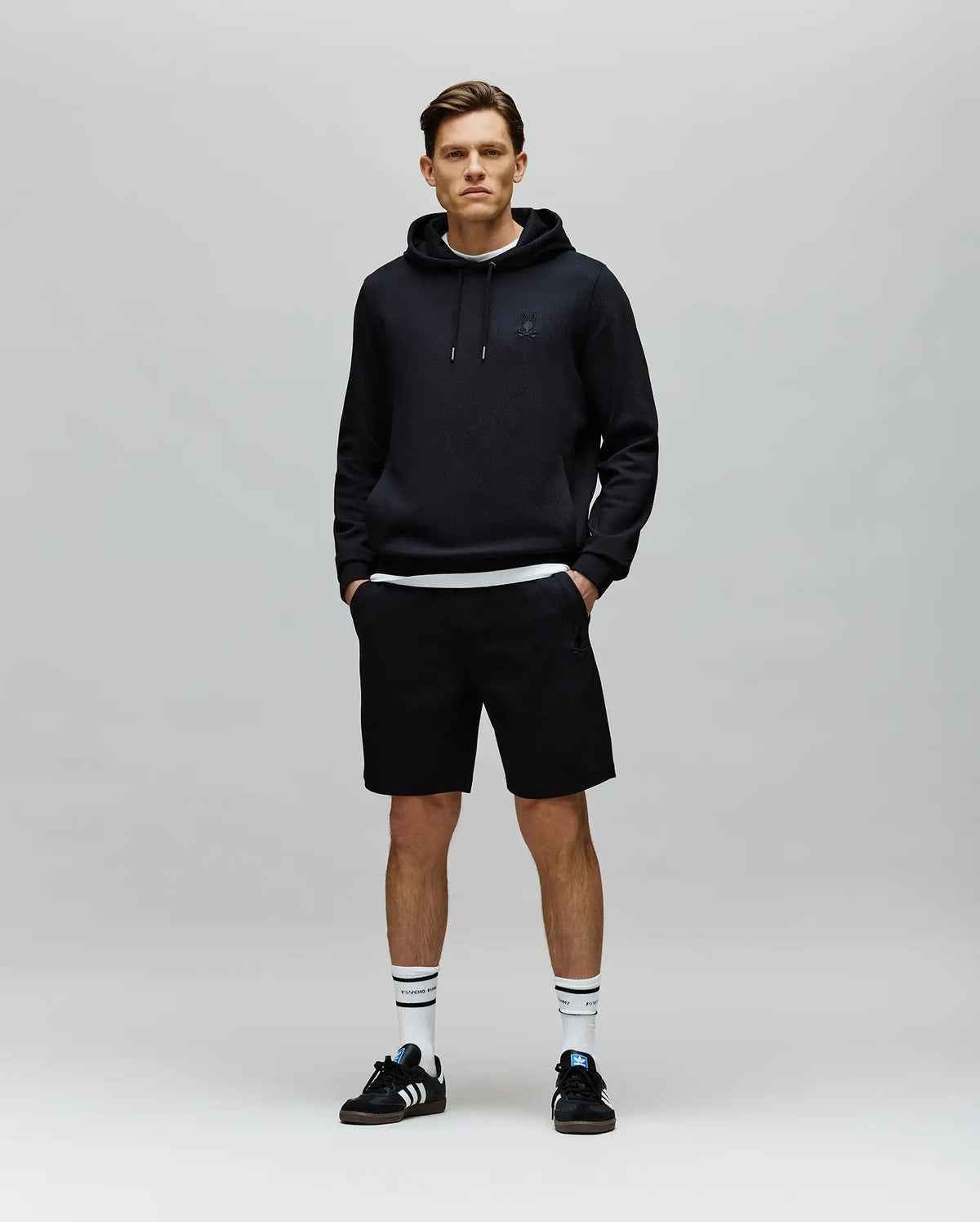 A young man stands against a plain gray background, wearing a black hoodie, Psycho Bunny MENS KAI SWEATSHORT - MB0900015, white socks with black stripes, and black Adidas sneakers. His hands are in his pockets as he faces the camera with a neutral expression.