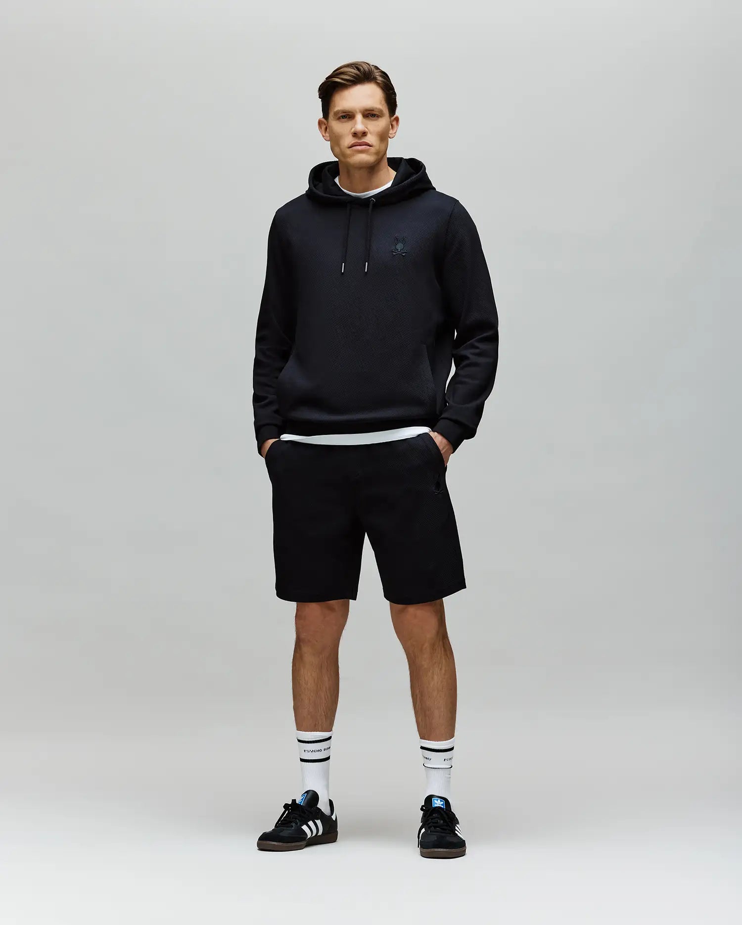 A young man stands against a plain gray background, wearing a black hoodie, Psycho Bunny MENS KAI SWEATSHORT - MB0900015, white socks with black stripes, and black Adidas sneakers. His hands are in his pockets as he faces the camera with a neutral expression.