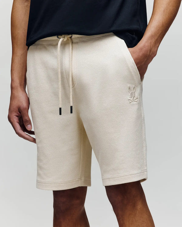A person wears Psycho Bunny MENS KAI SWEATSHORT - MB0900015 in beige twill texture, featuring a drawstring waistband, side pockets, and a small embroidered Bunny logo on the left leg. Their hand is in a pocket and they pair it with a black shirt.