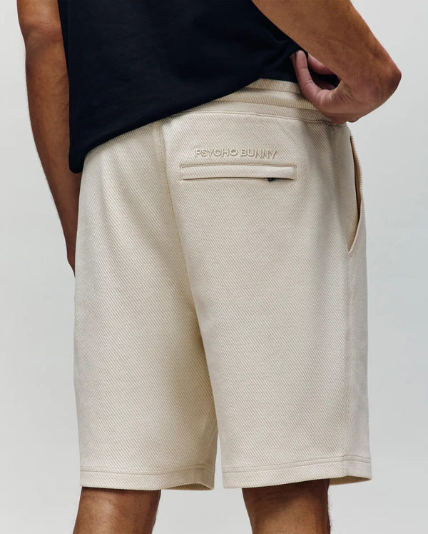 A person, seen from the back with one hand on their hip, wears light beige Psycho Bunny MENS KAI SWEATSHORT - MB0900015 featuring a twill-textured finish and an embroidered Bunny logo above the back pocket.