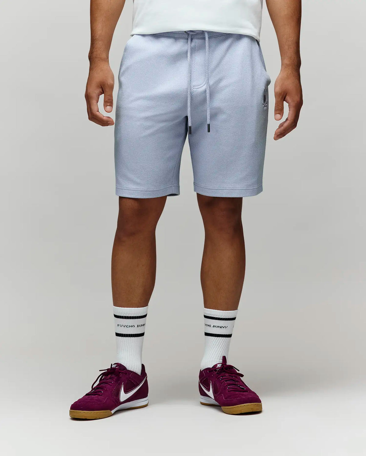A man stands against a plain light background, showing only his lower half. He wears Psycho Bunny’s MENS KAI SWEATSHORT - MB0900015 in light gray with the embroidered Bunny logo, paired with a white shirt, white crew socks, and maroon Nike sneakers.