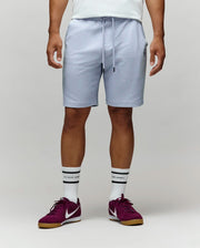 A man stands against a plain light background, showing only his lower half. He wears Psycho Bunny’s MENS KAI SWEATSHORT - MB0900015 in light gray with the embroidered Bunny logo, paired with a white shirt, white crew socks, and maroon Nike sneakers.