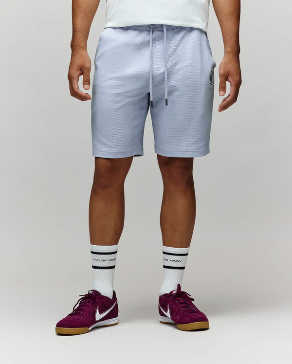 A man stands against a plain light background, showing only his lower half. He wears Psycho Bunny’s MENS KAI SWEATSHORT - MB0900015 in light gray with the embroidered Bunny logo, paired with a white shirt, white crew socks, and maroon Nike sneakers.