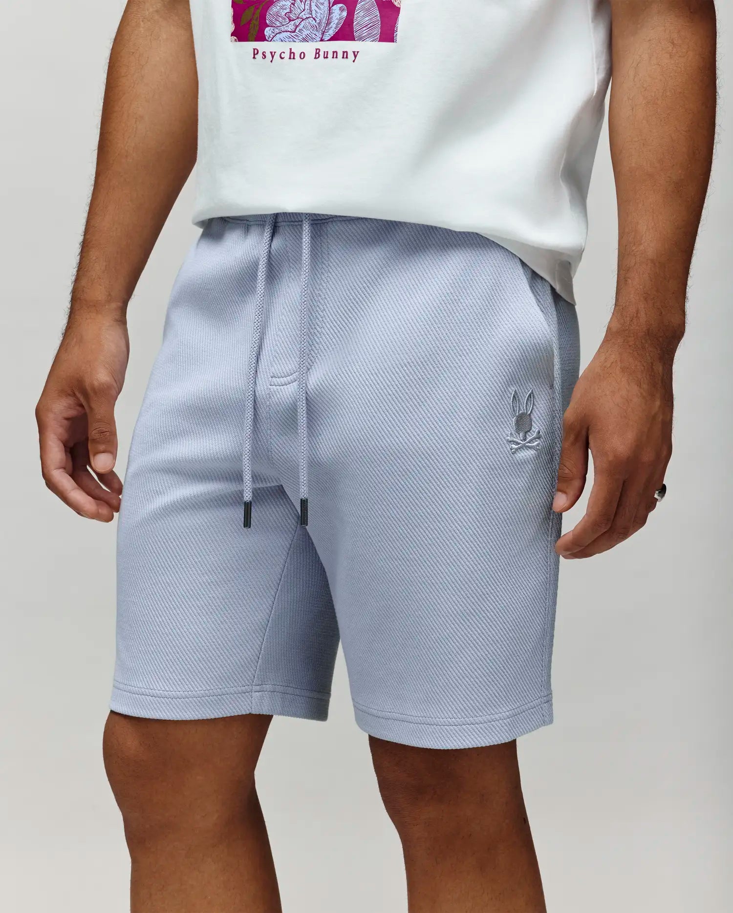 A person is shown from shoulders to knees wearing Psycho Bunny MENS KAI SWEATSHORT - MB0900015 in light gray, featuring drawstrings and an embroidered Bunny logo on the left thigh, paired with a white T-shirt.