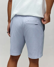 A person wearing Psycho Bunny’s MENS KAI SWEATSHORT – MB0900015 in light blue and a white T-shirt is seen from the back, with an embroidered Bunny logo above the pocket and their hand partially in the pocket.
