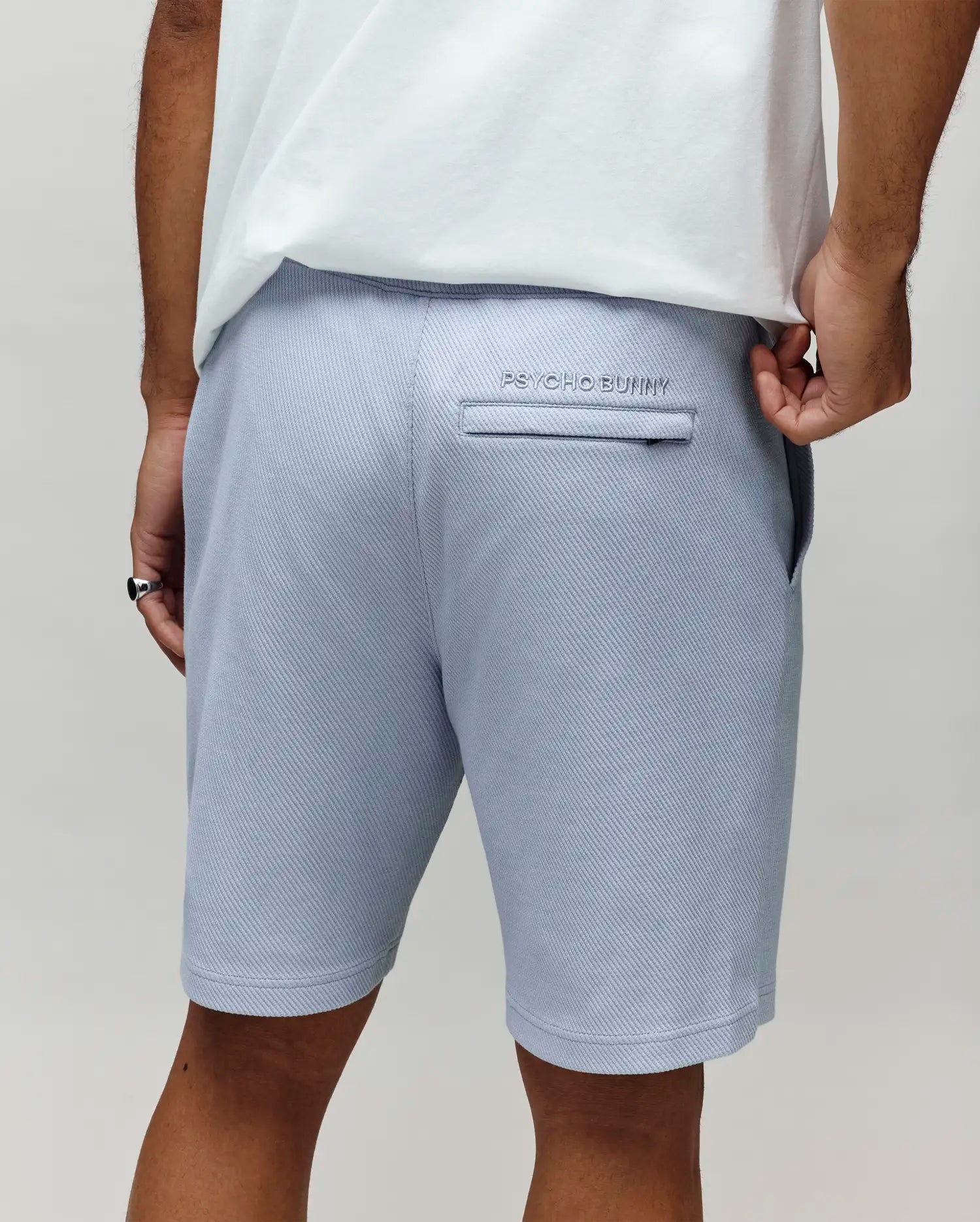 A person wearing Psycho Bunny’s MENS KAI SWEATSHORT – MB0900015 in light blue and a white T-shirt is seen from the back, with an embroidered Bunny logo above the pocket and their hand partially in the pocket.