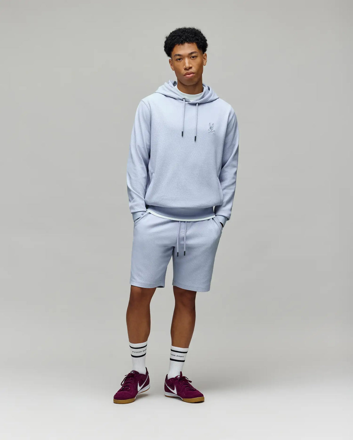 A young man stands against a plain background wearing Psycho Bunny’s MENS KAI SWEATSHORT (MB0900015), a light gray hoodie, white socks with black stripes, and maroon sneakers. His hands are in his pockets as he looks calmly at the camera.