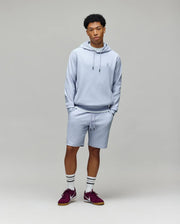 A young man stands against a plain background wearing Psycho Bunny’s MENS KAI SWEATSHORT (MB0900015), a light gray hoodie, white socks with black stripes, and maroon sneakers. His hands are in his pockets as he looks calmly at the camera.
