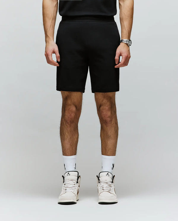 A person, shown from the shoulders down, wears Psycho Bunny MENS LAZSLO SWEATSHORT - MB0900016 in a stretch modal-cotton blend, paired with a black shirt, wristwatch, white logo socks, and white sneakers for an effortless casual look.