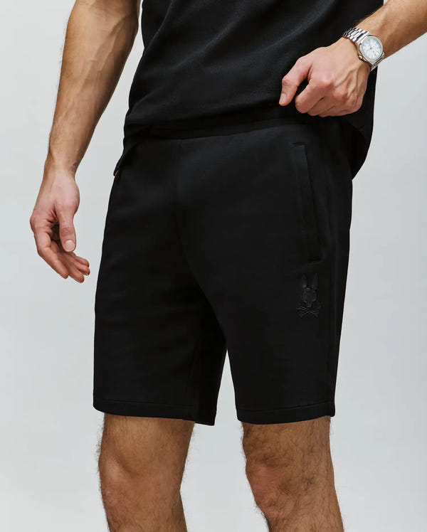 A person in a black T-shirt and Psycho Bunny MENS LAZSLO SWEATSHORT - MB0900016, made from stretch modal-cotton-blend with a subtle skull and bunny logo on the left leg, stands against a plain backdrop, sporting a watch for cool, casual style.