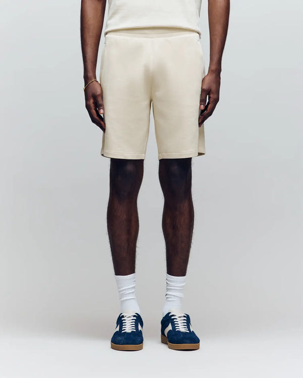 A person faces forward against a plain light background, wearing Psycho Bunny MENS LAZSLO SWEATSHORT - MB0900016 in cream, a matching stretch modal-cotton-blend t-shirt, white socks, and blue sneakers with brown soles.