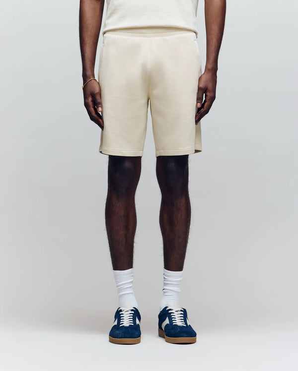 A person faces forward against a plain light background, wearing Psycho Bunny MENS LAZSLO SWEATSHORT - MB0900016 in cream, a matching stretch modal-cotton-blend t-shirt, white socks, and blue sneakers with brown soles.