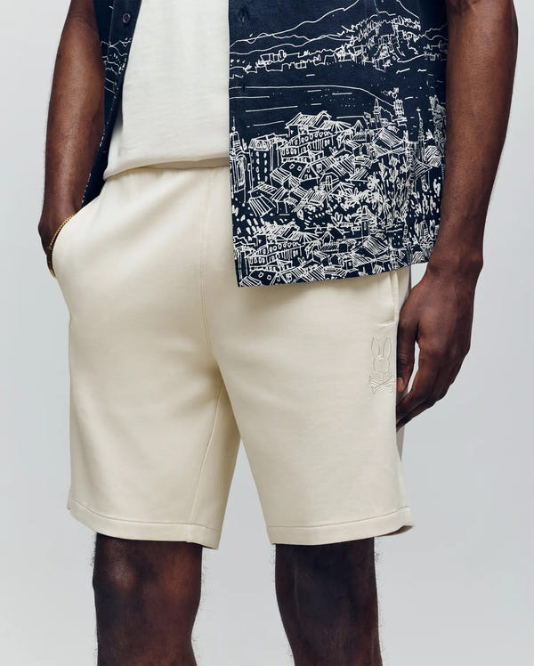 A person wears Psycho Bunny’s MENS LAZSLO SWEATSHORT - MB0900016 in a stretch modal-cotton blend, paired with a white shirt and a black-and-white patterned button-up featuring an embroidered Bunny, standing with one hand in their pocket.