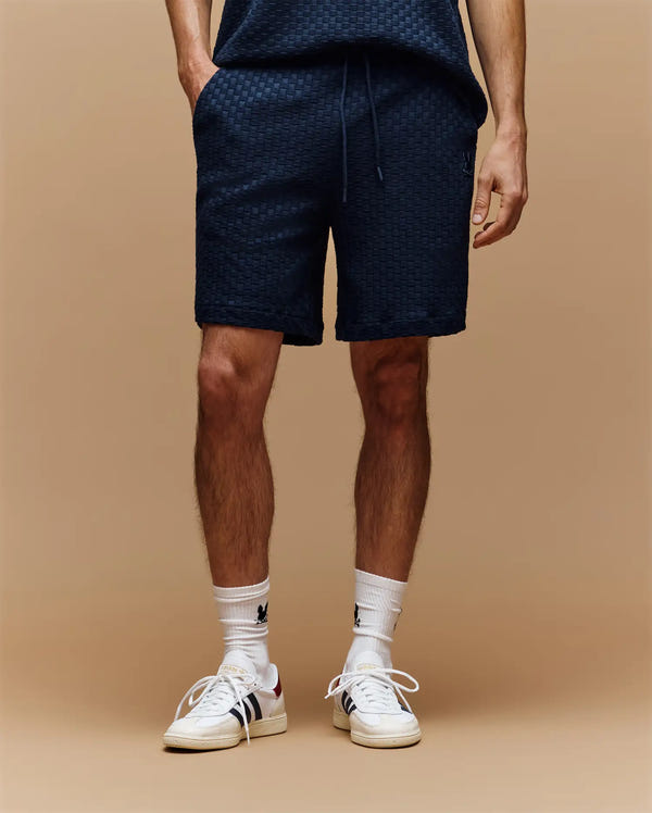 Against a beige background, only the lower half is shown, featuring Psycho Bunny MENS KAIRO CROCHET SHORT MB0900026 in navy cotton, paired with white crew socks with black embroidery and white sneakers with beige soles.