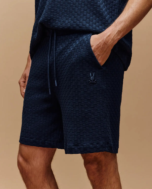 A man stands against a beige background, wearing Psycho Bunny's MENS KAIRO CROCHET SHORT - MB0900026 in textured navy cotton, one hand in the pocket and a small embroidered logo on the left leg.